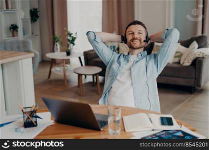 Happy male freelancer enjoying his remote job while using headset with microphone and laptop, sitting at his cozy home office and holding hands behind head. Freelance and distant work concept. Smiling young Caucasian man sitting at the table in living room