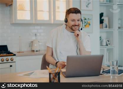 Happy male freelancer in headset sitting at kitchen table and smiling while having video call on laptop and working remotely. Online study and freelance job concept. Smiling guy holding hand on chin and looking at laptop screen