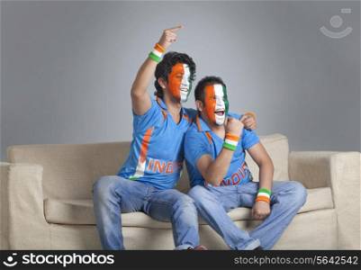 Happy male friends with face painted in Indian tricolor cheering while sitting on sofa