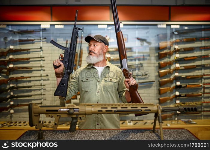 Happy male hunter with two rifles in gun store. Weapon shop interior, ammo and ammunition assortment, firearms choice, shooting hobby and lifestyle. Happy male hunter with two rifles in gun store