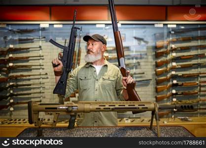 Happy male hunter with two rifles in gun store. Weapon shop interior, ammo and ammunition assortment, firearms choice, shooting hobby and lifestyle. Happy male hunter with two rifles in gun store