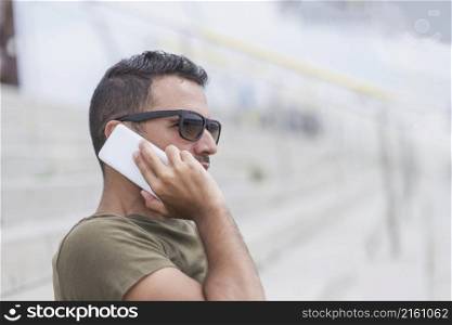Happy male in sunglasses standing outdoors, talking by smartphone