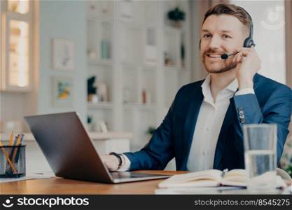 Happy male manager in suit wearing headphones and looking while working with laptop and talking online, businessman having online meeting while working remotely at home. Distant work concept. Smiling guy using headset and laptop while chatting on Internet at home