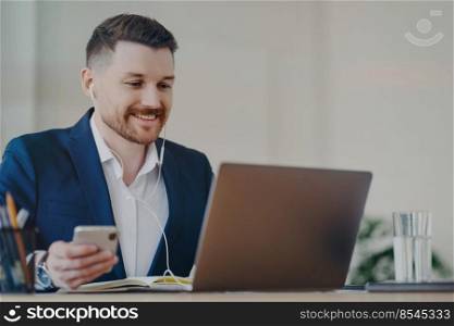 Happy male office worker uses modern technologies poses at desktop holds mobile phone makes video call via laptop wears earphones dressed formally has glad expression watches business webinar