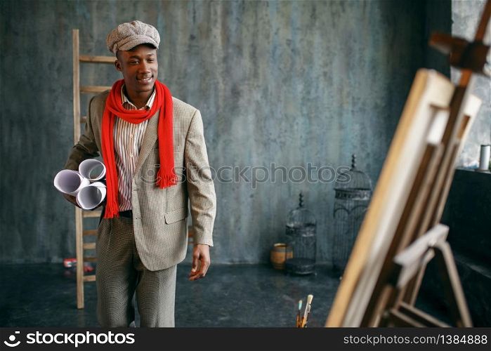 Happy male painter with rolls of canvases poses at the easel in art studio. Artist near his workplace, creative master works in workshop. Male painter with rolls of canvases in studio