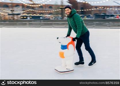 Happy male stands on ice ring on skates, leans on special aid for skating, has joy, poses at camera with happy expression. Unexperienced skater practices skating. Smiling man has free time enjoy hobby