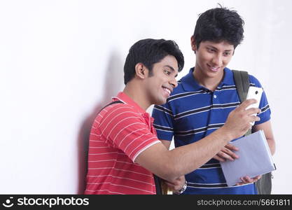 Happy male university students using mobile phone against wall