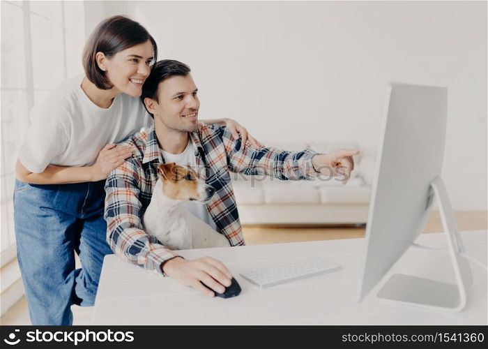 Happy man and his girlfriend consult about future project, point into monitor, pose at workplace together with dog in home interior, watch online tutorial video, spend time in modern apartment