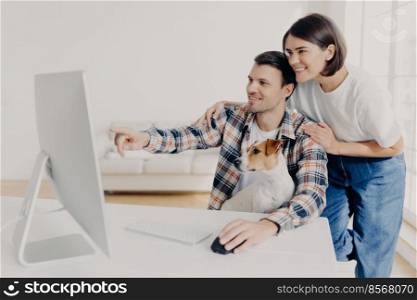 Happy man and his girlfriend consult about future project, point into monitor, pose at workplace together with dog in home interior, watch online tutorial video, spend time in modern apartment