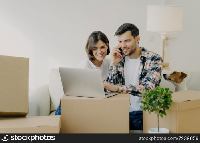 Happy man browses laptop in new flat, calls via smartphone, move in new apartment together with wife, sit on comfortable sofa, surrounded with cardboard boxes in living room, small dog near.