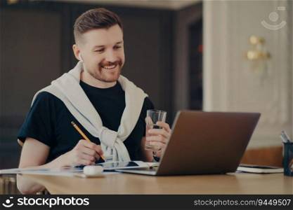 Happy man freelancer/entrepreneur, casual wear, laptop surfing, holding water glass, working online from home. Freelance concept.