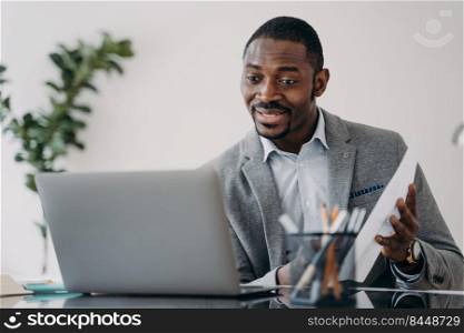Happy man has video call on laptop and pointing to data papers. Freelancer is working online at home office. Remote african american teacher or executive sitting in front of camera.. Happy man has video call on laptop and pointing to data papers. Remote african executive.