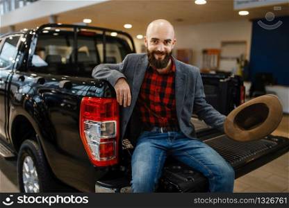 Happy man poses in the back of new pickup truck in car dealership. Customer in vehicle showroom, male person buying transport, auto dealer business. Happy man poses in the back of new pickup truck