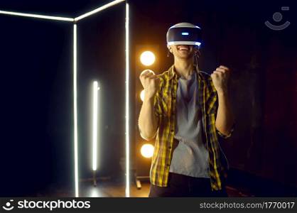 Happy man using virtual reality headset and gamepad in luminous cube, front view. Dark playing club interior, spotlight on background, VR technology with 3D vision. Happy man using virtual reality headset