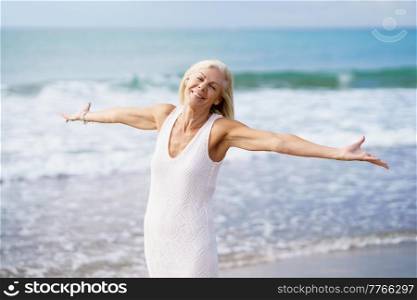 Happy mature woman opening her arms on a tropical beach, spending her leisure time. Elderly female enjoying her retirement at a seaside retreat.. Happy mature woman opening her arms on the beach, spending her leisure time, enjoying her free time