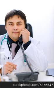 Happy medical doctor sitting at office table and speaking phone&#xA;