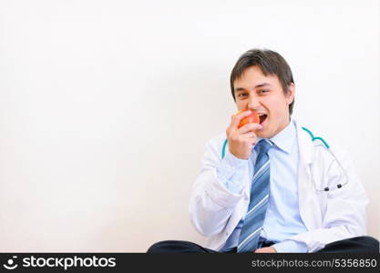 Happy medical doctor sitting on floor and eating apple