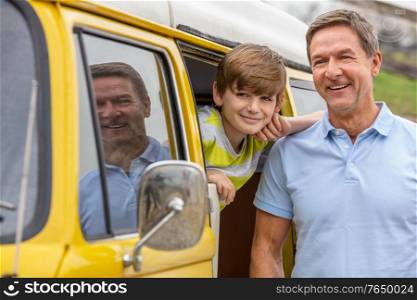 Happy middle aged man and boy male child, father and son together leaning out of a camper van bus in summer