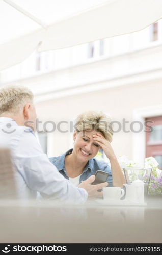 Happy middle-aged woman using mobile phone with man at sidewalk cafe