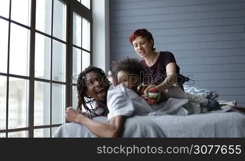 Happy mixed family relaxing in the bedroom at home. Smiling african father with dreadlocks lying on bed, caucasian mother putting their mixed race toddler son on daddy&acute;s back and tickling her boy. Joyful family with child lounging at home interior.