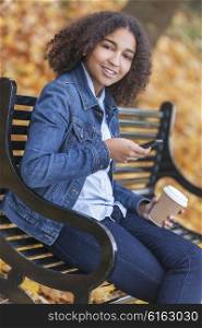Happy mixed race African American girl teenager female young woman drinking takeaway coffee &amp; texting on cell phone outside sitting on park bench in autumn or fall