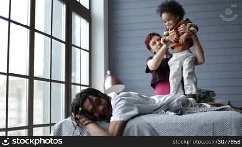 Happy mixed race family spending good time together in the bedroom at home. Handsome african dad with dreadlocks relaxing on the bed with his cute joyful toddler son lying on his back while caucasian playful mother tickling her sweet child.