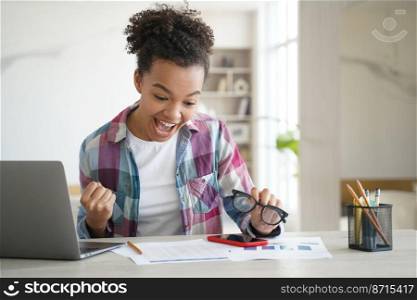 Happy mixed race girl student got good exam scores makes yes gesture, screaming, sitting at desk with laptop. Excited schoolgirl celebrates great news, university admission, personal achievement.. Overjoyed mixed race girl student celebrates good exam scores, makes yes gesture, screaming at desk