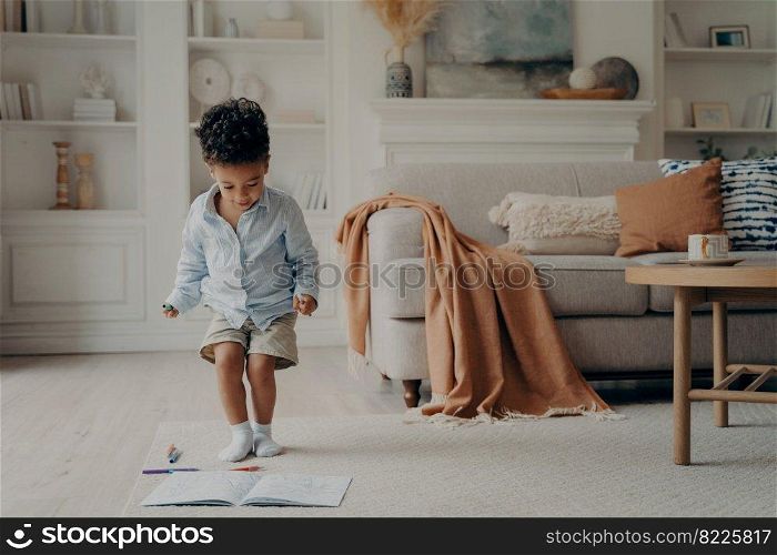 Happy mixed race little boy enjoying playing alone, drawing with pencils on warm carpeted floor, cute smiling african american kid having fun at home. Creative children activity indoor concept. Happy mixed race little boy enjoying playing alone at home, having fun in living room