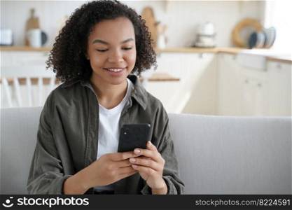 Happy mixed race teen girl holding smartphone uses mobile apps, shopping online, enjoying web surfing on internet. Smiling young lady with afro hair communicating in social networks at home.. Happy mixed race teen girl holding smartphone uses mobile apps, chatting or shopping online at home
