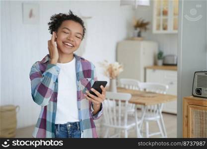Happy mixed race teen girl in earphones holds smartphone resting, using modern mobile apps, listening music online at home. Smiling biracial lady wearing earbud enjoying musical playlist indoors.. Happy mixed race girl in earphone holds smartphone using modern mobile apps, listening music at home