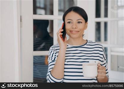 Happy mixed race woman is talking on smartphone. Lady is standing at the balcony with cup of coffee and smiling. Woman in striped sailor style shirt. Coffee break, remote work online from home.. Happy woman is talking on smartphone. Lady standing at the balcony with cup of coffee and smiling.
