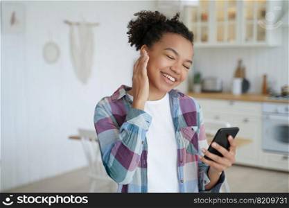 Happy modern young mixed race girl listening to music, holding smartphone, using musical apps, standing at home. Smiling biracial teen lady with afro hairstyle enjoying melody sound indoors.. Modern young mixed race girl listening to music, holding smartphone, using musical apps at home