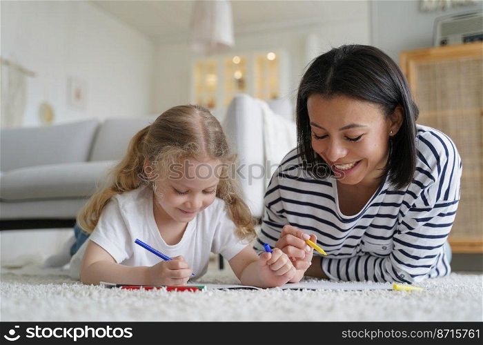 Happy mom and adopted daughter drawing together, lying on warm floor at home. Mother or babysitter helps preschooler kid girl to paint with colorful pencils. Adoption, children’s creative leisure.. Happy mom, adopted daughter drawing together, lying on floor. Adoption, children’s creative leisure