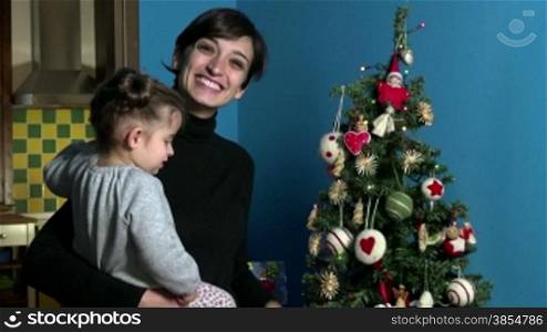 Happy mom and child, woman holding cute daughter, mother and baby looking at Christmas tree. People, children, holidays at home, xmas, lifestyle. Portrait of woman smiling at camera. 1 of 5
