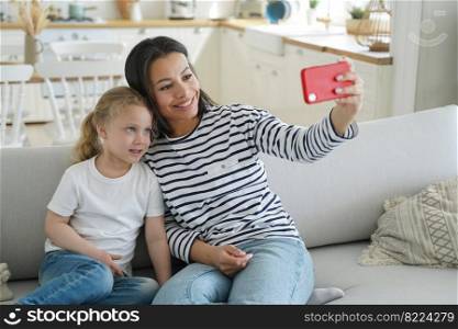 Happy mom and little daughter takes home selfie together, sitting on sofa, holding smartphone, looking at phone screen. Smiling mother and kid girl makes video call, chatting online, using mobile app.. Happy mom, little daughter takes selfie together or makes video call, holding phone, sitting on sofa
