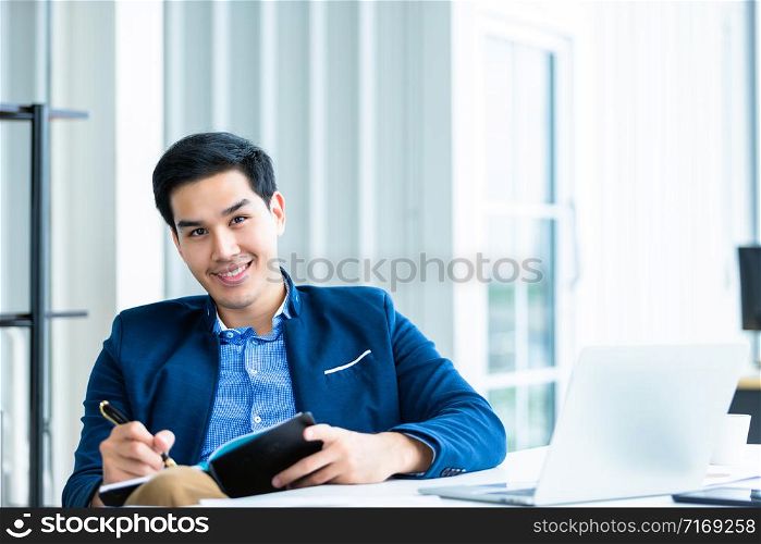 Happy mood a cheerful of asian young businessman working with making notes in a notebook and laptop computer In the office room background.