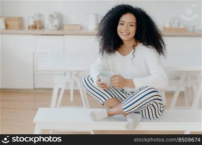 Happy morning concept. Photo of cheerful curly African American woman sits in lotus pose at white bench, sips tasty aromatic drink, feels relaxed, poses against kitchen interior, smiles broadly