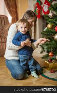 Happy mother and 10 months old baby boy decorating Christmas tree at house