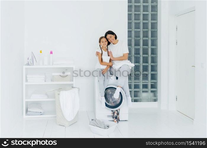 Happy mother and daughter have fun, embrace and express love to each other, dressed in casual wear, sit on washer, prepare for washing, being in white spacious laundry room, basin with dirty clothes