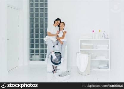 Happy mother and daughter have fun, embrace and express love to each other, dressed in casual wear, sit on washer, prepare for washing, being in white spacious laundry room, basin with dirty clothes