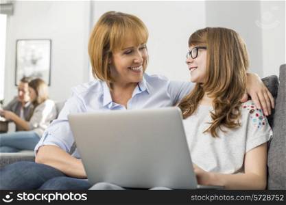 Happy mother and daughter with laptop while family sitting in background at home
