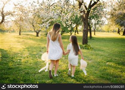Happy mother and her daughter in white dresses run through the spring garden at sunset