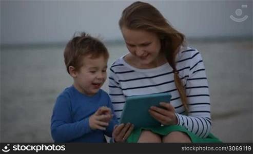 Happy mother and her little son enjoying playing on tablet computer by the sea