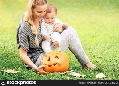 Happy mother and little baby boy outdoors, sitting on green grass field with orange carved pumpkin decorative toy, preparation to Halloween holiday