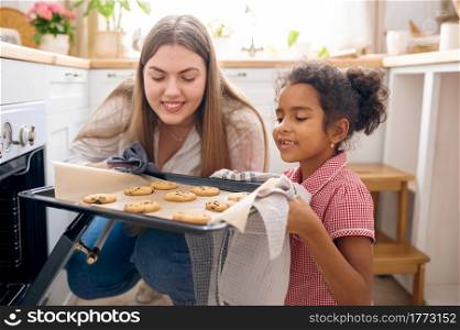 Happy mother and little kid cooking cakes in oven on breakfast. Smiling family on the kitchen in the morning. Mom feeds female child, good relationship. Mother and kid cooking cakes in oven on breakfast