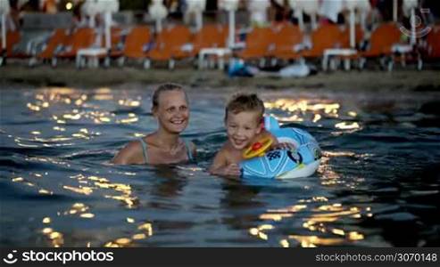 Happy mother and little son swimming in sea near the shore in the evening. Boy using rubber ring made as car