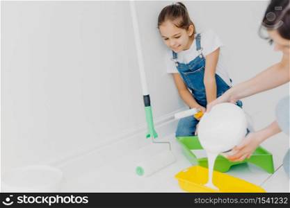 Happy mother and small daughter make repairing at their home, going to paint walls, pour white paint in tray, poses in spacious room, busy with house renovation, redecoration or refurbishment
