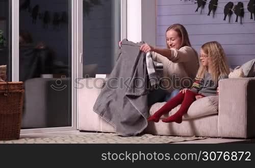Happy mother and smiling child wrapping in blanket on the sofa in living room during winter. Young mom hugging and cuddling her daughter on the couch. Two are snuggled with blanket, girl holding teddy bear.