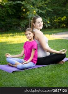 Happy mother and smiling daughter practicing yoga and sitting back to back at park