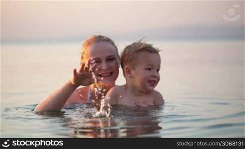Happy mother and young child waving goodbye while immerse in the sea water during sunset in the beach